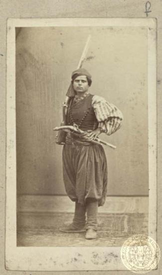 Νεαρός άνδρας με τη χαρακτηριστική νησιώτικη ενδυμασία οπλισμένος με τουφέκι και κουμπούρι περασμένο στη ζώνη.