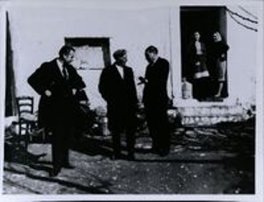 Τρείς άνδρες να συζητάνε ενώ παράλληλα δύο γυναίκες τους παρακολουθούν από την πόρτα του σπιτιού.