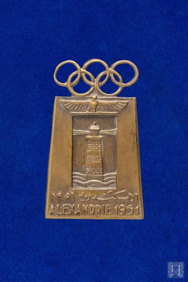 Αναμνηστική καρφίτσα με σχήμα τον Φάρο της Αλεξάνδρειας, με παράσταση πτηνό και το σήμα των Ολυμπιακών Αγώνων και επιγραφή ALEXANDRIE 1951