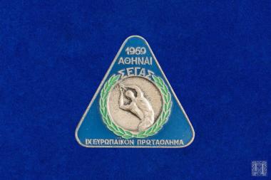 Αναμνηστική  καρφίτσα τριγωνικού σχήματος με στρογγυλεμένα άκρα, αναμνηστική καρφίτσα με μπλε και πράσινα σμάλτα και την επιγραφή 1969 ΑΘΗΝΑΙ ΣΕΓΑΣ IX ΕΥΡΩΠΑΪΚΟΝ ΠΡΩΤΑΘΛΗΜΑ Στην οπίσθια όψη φέρει την καρφίτσα και υποδοχή για κορδέλα.