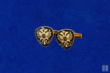 Δύο μανικετόκουμπα από κράμα χαλκού τριγωνικού σχήματος με στρογγυλεμένες γωνίες. Φέρει θυρεό με δικέφαλο αετό σε μαύρο σμάλτο