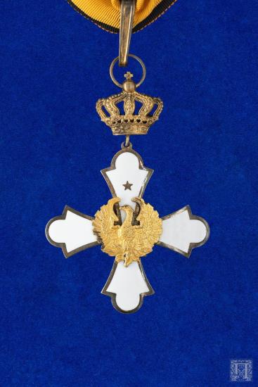 Μεγαλόσταυρος με κίτρινη γκρο κορδέλα, με διακόσμηση σε λευκό σμάλτο και κεντρικά στοιχεία στην εμπρόσθια όψη έναν Φοίνικα και στην οπίσθια, το μονόγραμμα Π (Παύλος) και την κορώνα από πάνω του. Ανάμεσα στον σταυρό και την κορδέλα υπάρχει ως ενδιάμεσο συνδετικό διακοσμητικό μέσο, μία κορώνα από κράμα χαλκού με μεγάλο κρίκο.