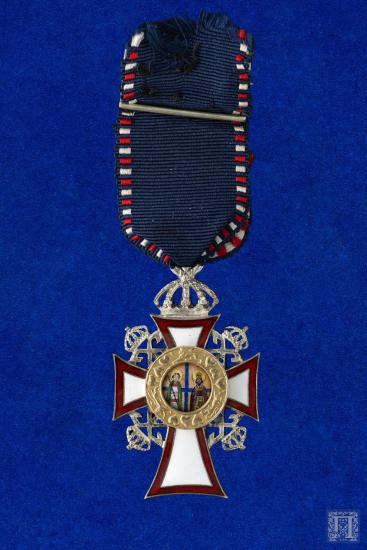 Μεγαλόσταυρος σε  μπλε σκούρα γκρο κορδέλα με ακτίνες από κόκκινο και λευκό σμάλτο, με διακόσμηση ένα καλλιγραφικό Χ με μία κορώνα ανάμεσά τους ανά δύο και στο κέντρο μεταλλικό διακοσμητικό. Το τελευταίο στη μία όψη απεικονίζει δύο Αγίους και στην άλλη, ένα οικόσημο με στέμμα