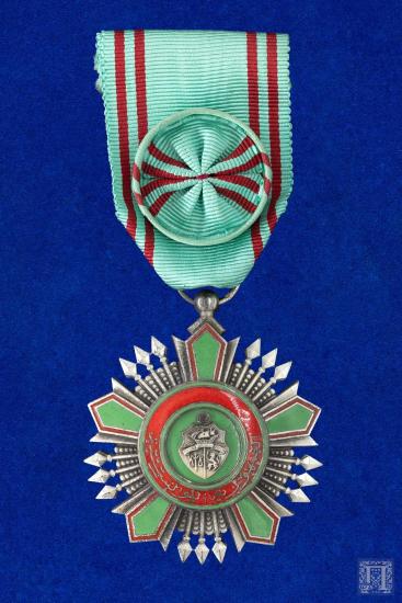 Μετάλλιο με πράσινη γκρο κορδέλα με λεπτές κόκκινες ρίγες, σε πράσινο και κόκκινο σμάλτο με οικόσημο στο κέντρο και περιμετρικά, επιγραφή στα αραβικά