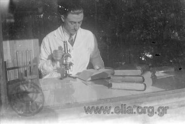 Γιατρός ή χημικός σε εργαστήριο. Δίπλα του, στον πάγκο, μικροσκόπιο και δοκιμαστικοί σωλήνες.