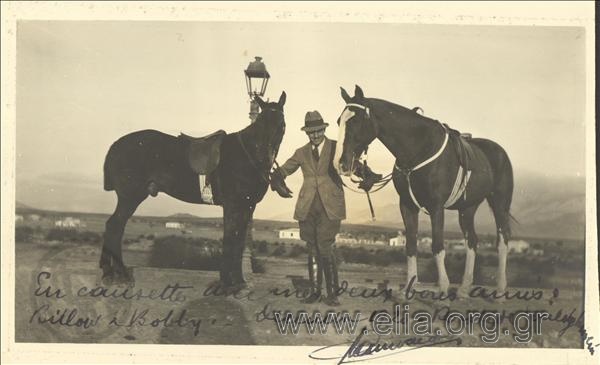 Άνδρας κρατά από τα χαλινάρια δύο άλογα. Στο βάθος οικισμός (περιοχή Βουλιαγμένης)