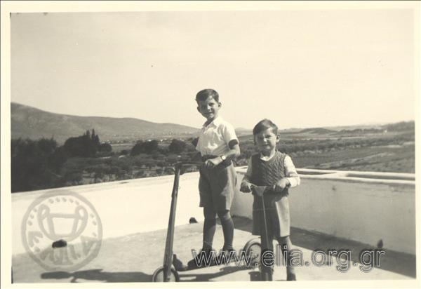 Small children Marinos and Giorgos I. Geroulanos on a veranda with a scooter