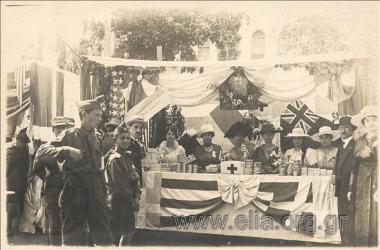Stand with products of Greek  workshops made in Fanari in international bazzar (?) probably in Smyrna. Ladies-Salesgirls pose behind the stand. A photograph of Eleftherios Venizelos decorated with flowers hangs on the wall.