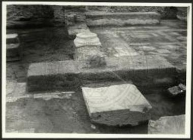 Μαρμαρίνη πλίνθος και πεσσός εις σχήμα Τ. Όπισθεν του πεσσού ο στηλοβάτης του τριβήλου.