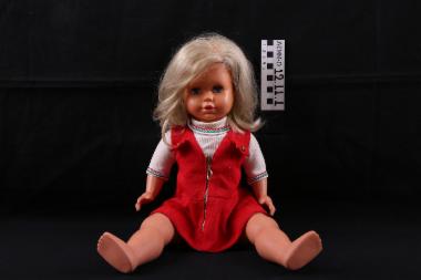 Κούκλα - κούκλα πλαστική (κορίτσι) με μηχανισμό, πιθανόν, παραγωγής φωνής, κινούμενα μέλη, μαλλιά ξανθά, μάτια ανοιγοκλειόμενα γαλανά. Φέρει φόρεμα κόκκινο αμάνικο και μπλουζάκι λευκό. Αγορασμένη από τη Γερμανία