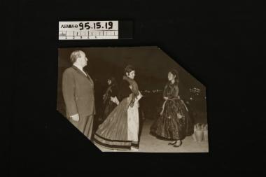 Φωτογραφία - φωτογραφία ασπρόμαυρη που απεικονίζει ηλικιωμένο άνδρα με κοστούμι που επιτηρεί νεαρές γυναίκες που χορεύουν, ντυμένες με παραδοσιακές φορεσιές