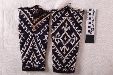 Κάλτσες (ζεύγος) - περικνημίδες μάλλινες πλεκτές σε χρώμα λευκό με μπλε και μαύρο κάμπο, με γεωμετρικά σχέδια, με κεντρικό μοτίβο ρόμβου που εσωκλείει σταυρόσχημο λευκό μοτίβο και έναν μικρό λευκό ρόμβο με σταυρό στο κέντρο. Σαρακατσάνικες