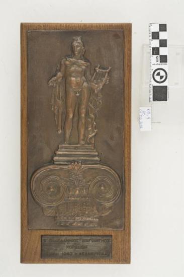 Τιμητική πλακέτα με ανάγλυφη απεικόνιση γυναικείας μορφής να κρατά αρχαία ελληνική μουσική λύρα και να στέκεται πάνω σε κιονόκρανο διακοσμημένο με ανάγλυφα φυτικά κοσμήματα