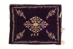 Eggplant violet velvet with gold embroidery in couching technique, spiraled threads and sequins, central floral ornament, corner motifs and foliate border, beige ribbon ties at both ends.