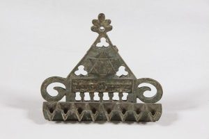Oil lamp with eight lights used for the holiday of Hanukkah, cast and cut brass, triangular back wall with Star of David, row of cut horseshoe arches and inscription in low relief 'The commandment is a candle and the Torah is light', missing Shamash and wick container.