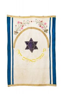 Torah ark curtain, white silk satin, trimmed on both sides with blue silk satin stripes, on top and bottom with woven gold braids, multicoloured embroidered bouquets, yellow embroidered inscription adapted from Psalms, central blue velvet dedication panel in shape of the Star of David, dedicated by Esther, wife of Hayim Bar Hayim Kabeli, Ioannina.