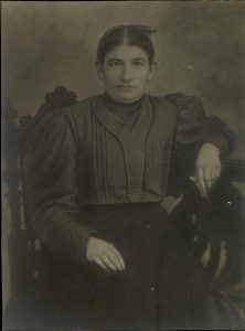 Woman in high-necked dark dress, hair braided and pinned upm sitting on a wooden sofa.