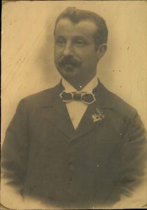 Man with moustache wearing dark jacke with flower in buttonhole, shirt with shirt collar and bow tie.