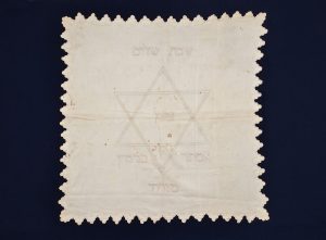 White cotton tablecloth for Kiddush, with scalloped edge, embroidered with white thread, central motif of Star of David inscribed with 'Zion', 'Shabbat Shalom' (above) and 'Esther Benjamin Mizrahi' (below).