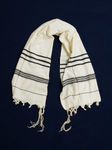 Prayer shawl, cream cotton with black stripes along the edge, white square corner reinforcements.