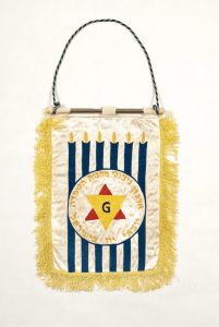 Banner of the Organization for Greek Survivors of Concentration Camps in Israel, machine-embroidered satin, motif of six lit blue candles with emblem of organization, edged with yellow fringe trim, from Israel.