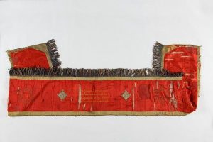 Bimah front cover, red silk, edged with gold braid and fringe trim, yellow embroidered inscription flanked by lozenge-shaped embroidery appliques and motifs of Star of David, dedicated by Zoya in memory of her husband Joseph Matathia Halevi, who died in 1901.