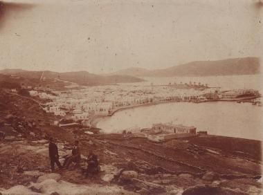 Άποψη της Χώρας Μυκόνου από τα βόρεια. Τρεις άνδρες σε βραχώδη λόφο πάνω από την πόλη.