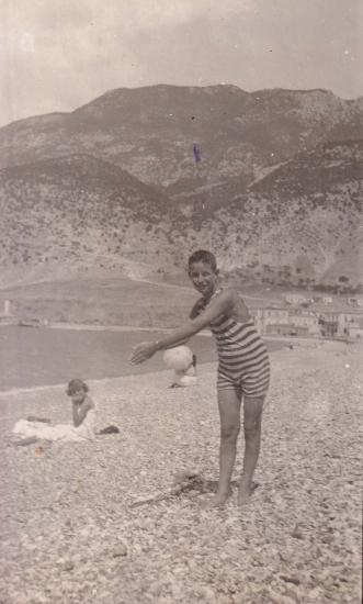 Αγόρι με το μαγιό του ποζάρει στην παραλία του Λουτρακίου, με τον οικισμό στο βάθος.