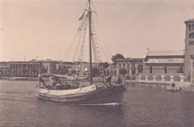 Καΐκι προσεγγίζει το λιμάνι της Ρόδου (Μανδράκι). Στα δεξιά, ο καθεδρικό ναός του Ευαγγελισμού της Θεοτόκου (πρώην καθολικός ναός του Αγίου Ιωάννη) και αριστερά το δικαστικό μέγαρο της Ρόδου.