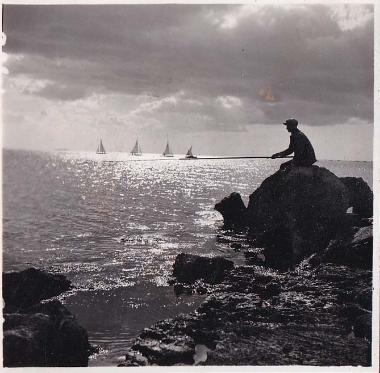 Άνδρας καθιμσένος σε βράχο ψαρεύει με καλάμι.