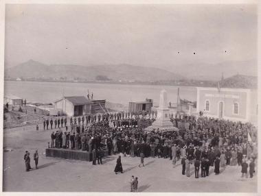 Κόσμος συγκεντρωμένος γύρω από το μνημείο των Φιλελλήνων στην ομώνυμη πλατεία του Ναυπλίου∙ στα δεξιά το κτήριο του Τελωνείου. Το μνημείο, στον τύπο του οβελίσκου, σχεδιάστηκε στο Παρίσι, κατασκευάστηκε στην Αθήνα από τον γλύπτη Ιωάννη Χαλδούπη και ανεγέρθηκε το 1903.