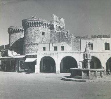Η πλατεία Ιπποκράτους (Παλαιά Αγορά) στην πόλη της Ρόδου, με τις μεσαιωνικές οχυρώσεις και την πύλη της Αγίας Αικατερίνης πίσω∙ κρήνη στα δεξιά.